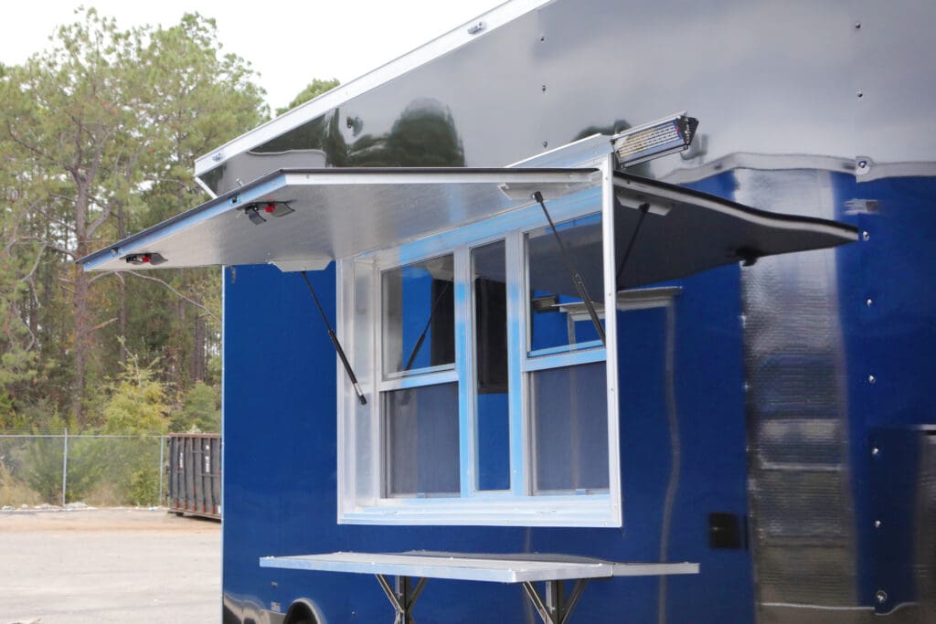 A blue food or concession trailer with a large open serving window and a pull-out counter below it.